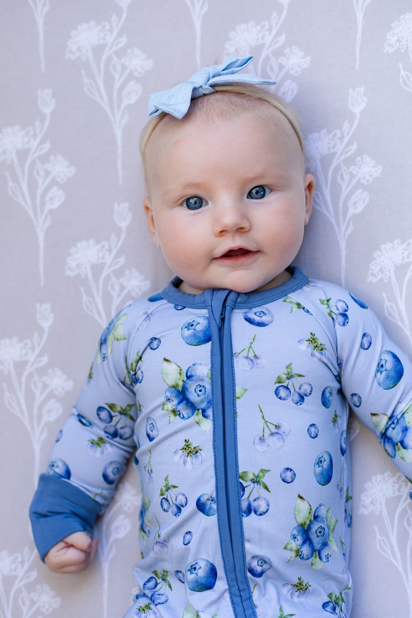 A baby wearing a light blue bamboo sleeper with blueberry print and a matching blue bow headband, lying on a floral-patterned light gray blanket.